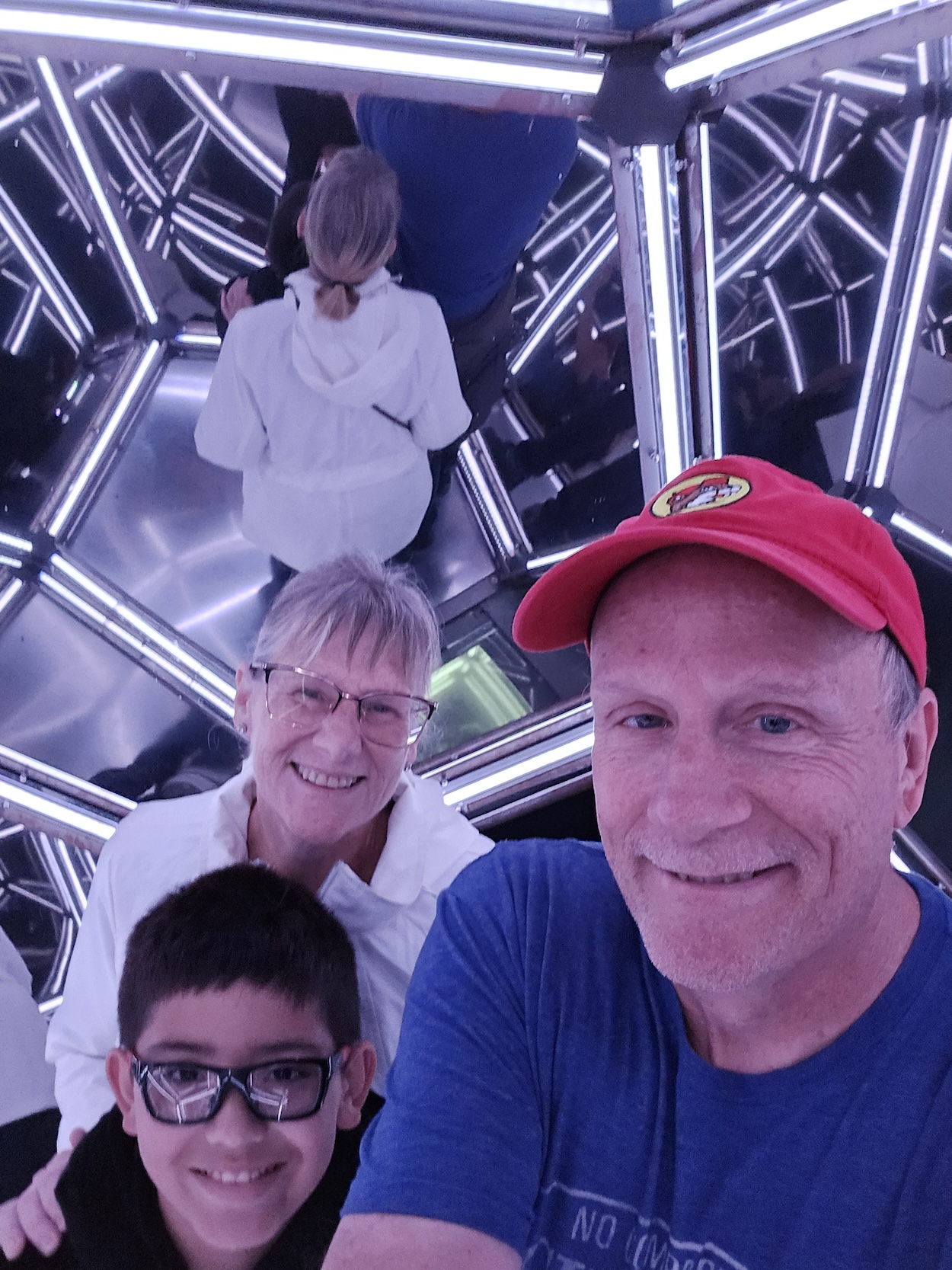 Three people smiling for a selfie inside a mirrored light installation with geometric reflections.
