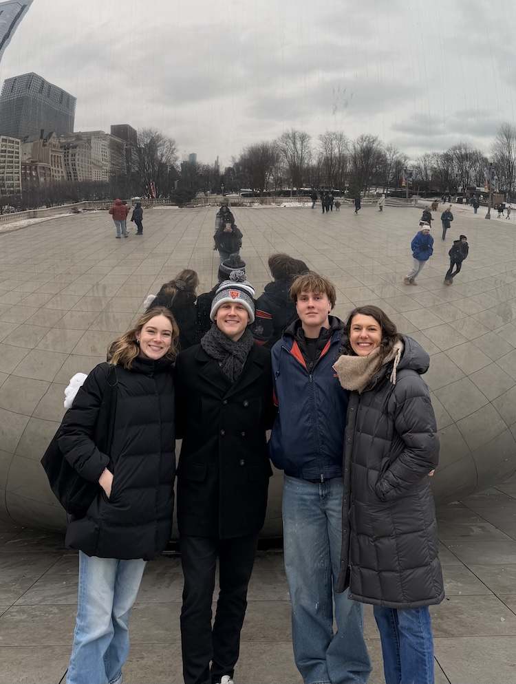 Four people in winter coats pose together in front of Cloud Gate in Chicago on an overcast day.