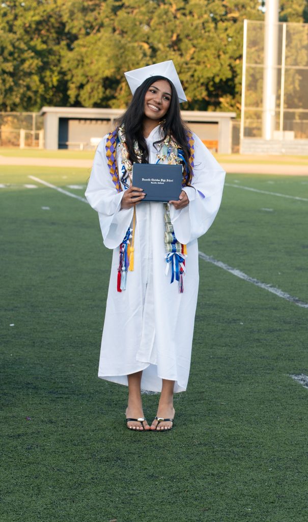Vacaville Christian High School graduate in a white cap and gown holding a diploma on the football field after commencement.