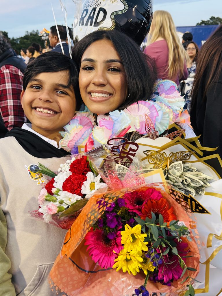 Graduate smiling with a young boy while holding colorful flower bouquets, a money bouquet, and graduation balloons during an outdoor celebration.