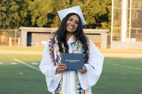 Vacaville Christian High School graduate in a white cap and gown holding a diploma on the football field after commencement.